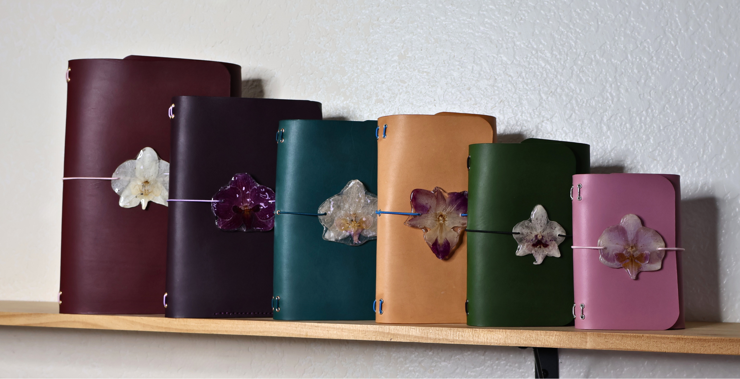 Set of colorful leather journals with floral charm on a shelf against a white wall.