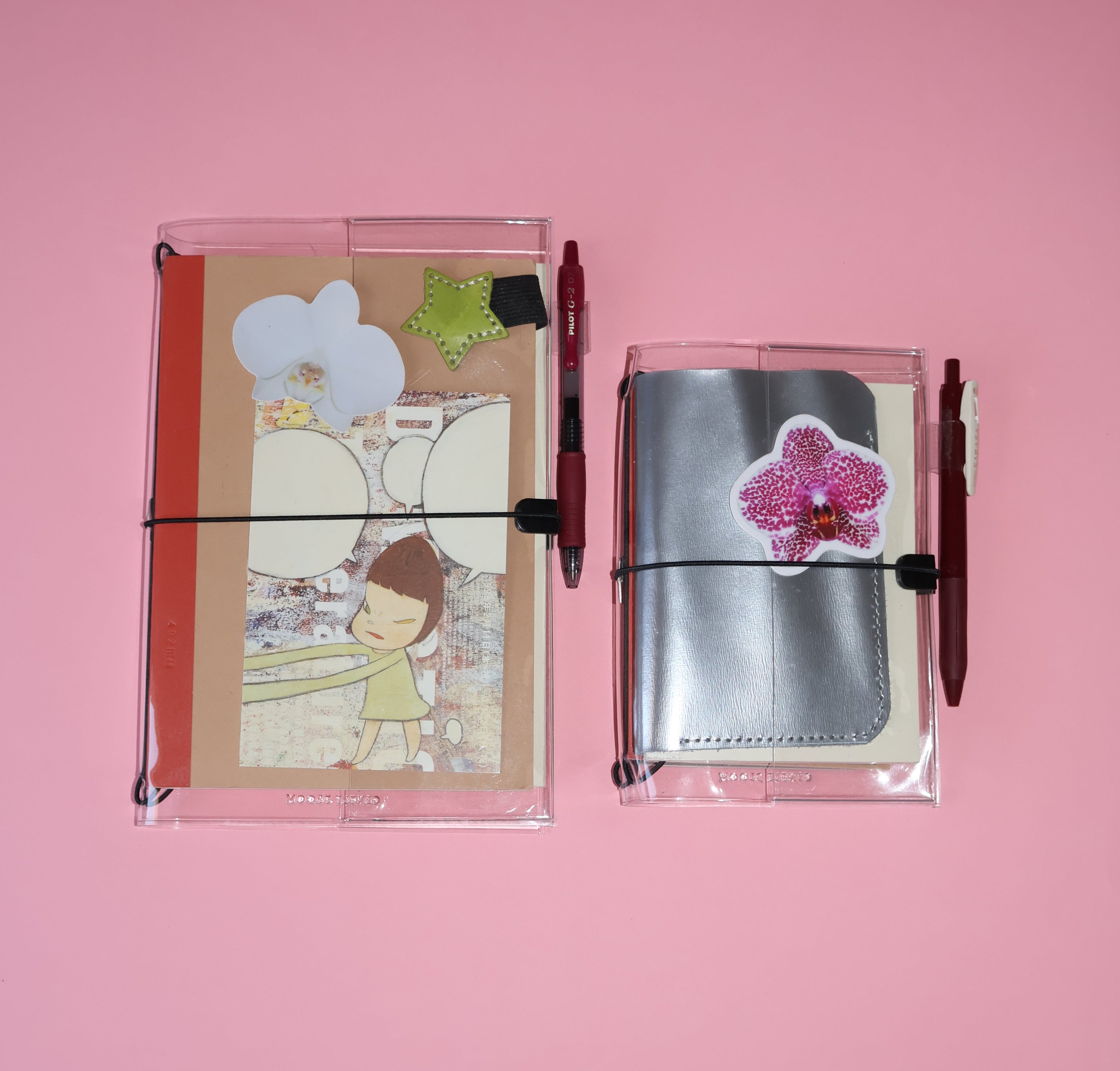 Two clear plastic pen pouches with elastic closures on a solid pink background. The left pouch contains a decorated card and a red pen, while the right pouch contains a silver notebook and a pink flower applique with a red pen.