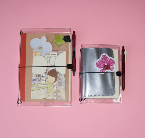 Two clear plastic pen pouches with elastic closures on a solid pink background. The left pouch contains a decorated card and a red pen, while the right pouch contains a silver notebook and a pink flower applique with a red pen.