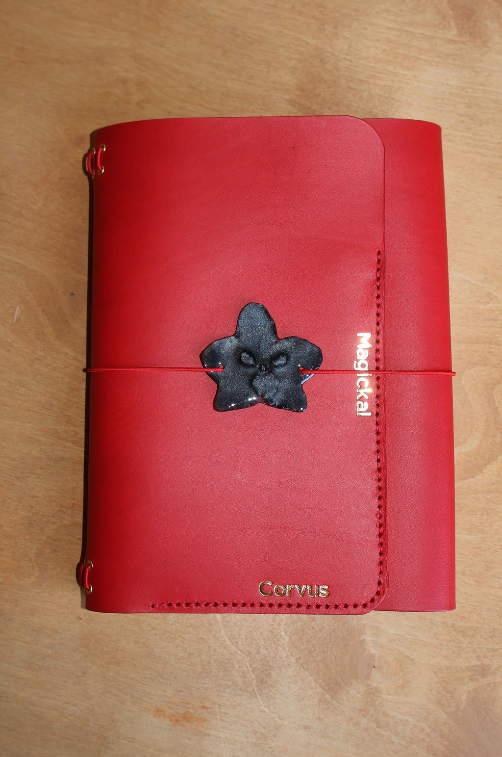 A red leather journal with a black flower-shaped metal clasp closure, laid flat on a light brown wooden surface. The cover features a vertical pocket with 'mochila' written in white, and 'Cinnabar' at the bottom.