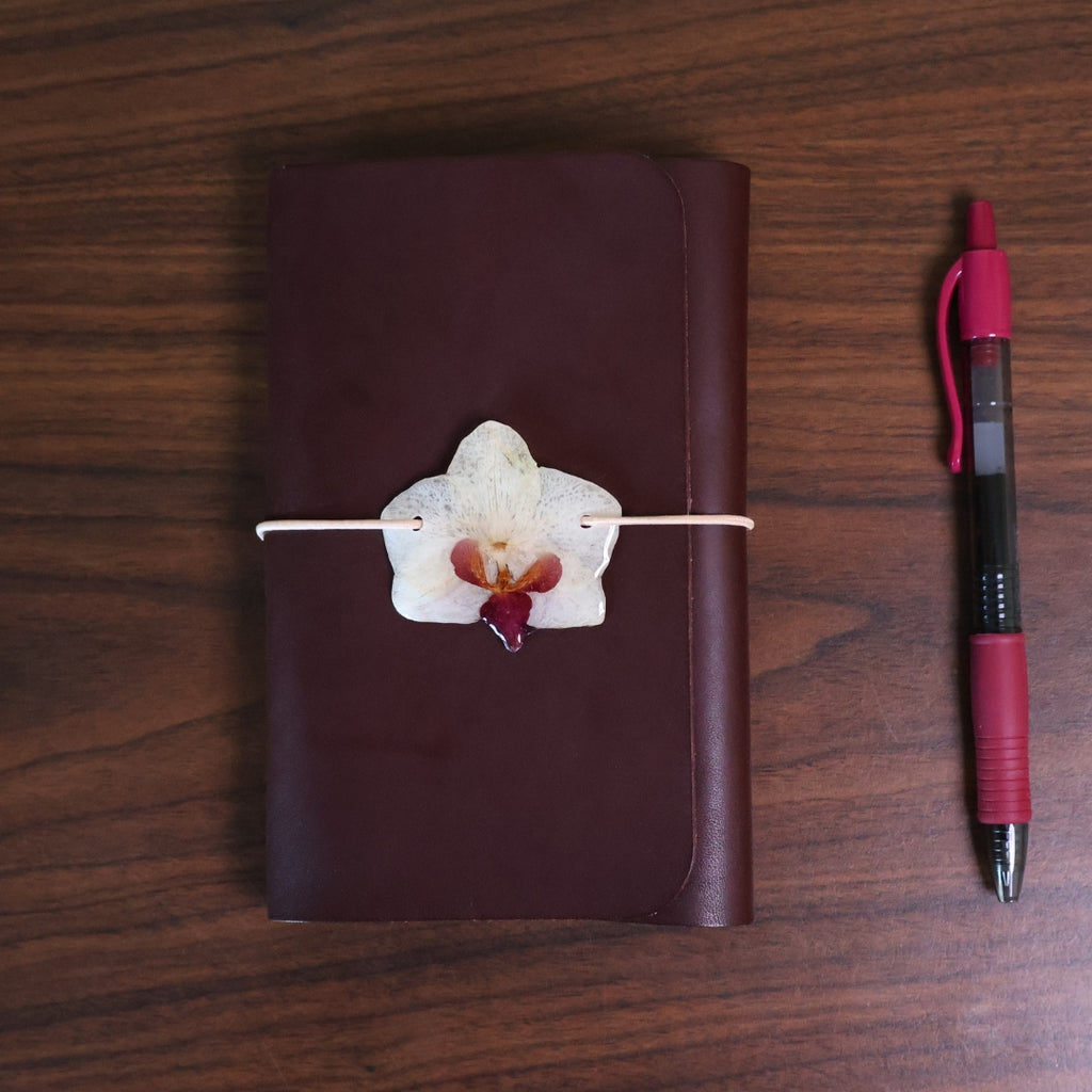 Brown leather notebook with a decorative orchid on a wooden surface next to a red pen.