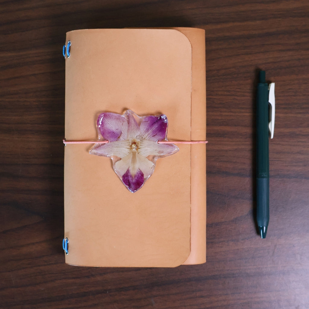 Brown leather notebook with a purple flower decoration on a wooden surface, accompanied by a black pen.