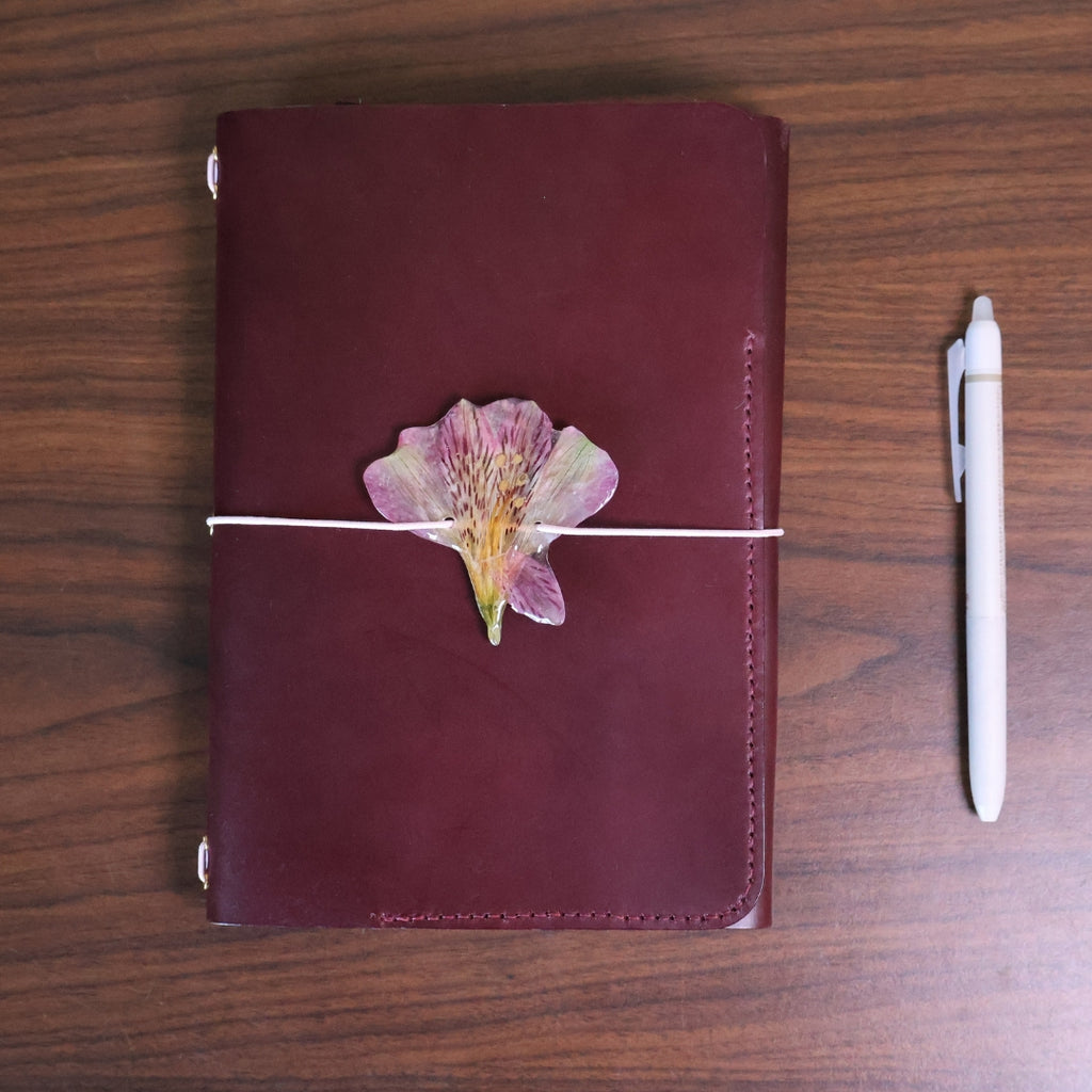 Maroon leather notebook with a pressed flower on a wooden surface next to a white pen.