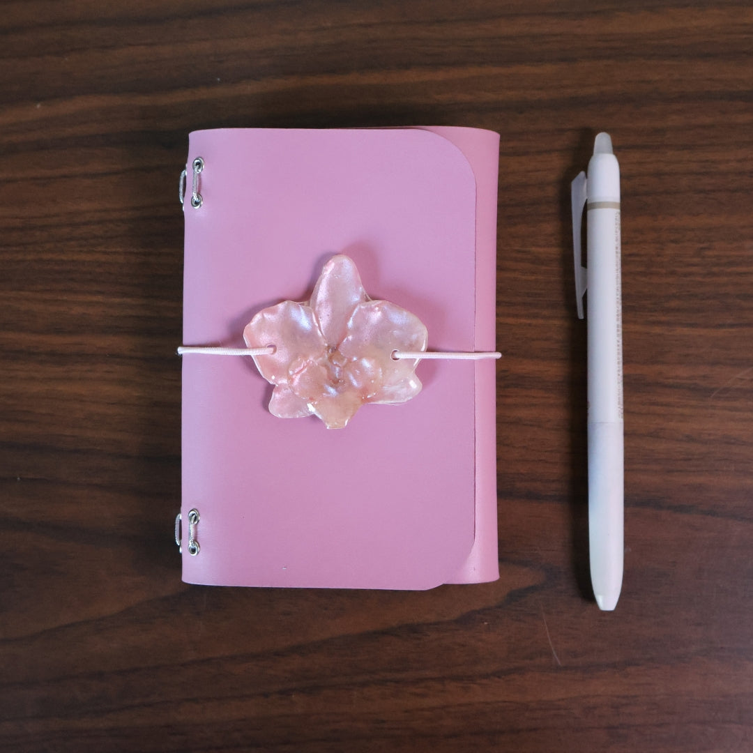 Pink notebook with floral design and white pen on wooden surface